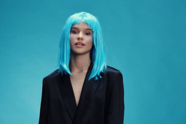 Mavi arka planda siyah takım elbiseli mavi saçlı bir kadın, mavi bir peruk takıyor ve kameradan uzağa bakıyor, çarpıcı ve yapışkan bir görsel karşıtlık oluşturuyor.