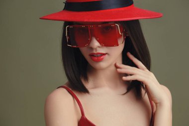 Kırmızı şapkalı, güneş gözlüklü, eli çenesinde poz veren, yana bakan kadın düşünceli bir şekilde bakıyor fotoğrafa gizem ve tarz katıyor.