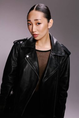 Asyalı kadın siyah deri ceket kamera grisi arka plan pozu veriyor. Kendine güvenen ve modaya uyan bir kadın için şık ve şık bir görünüş..