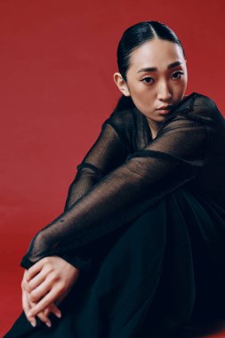 Kırmızı arka planda oturan, elleri göğsü ve gözleri kapalı, siyah elbiseli Asyalı kadın. şık ve zarif bir moda imajı..