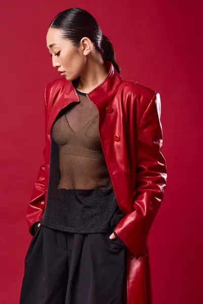 stock image Fashionable asian brunette in red leather jacket and black pants posing with hands on hips and eyes closed against a red background.