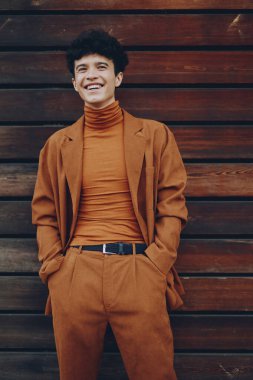 Turuncu balıkçı yaka ve takım elbise giyen neşeli genç bir adam elleri cebinde, kırsal bir ahşap zemine karşı güvenli bir şekilde duruyor.