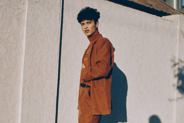 Модный молодой мужчина-модель в коричневом наряде стоит напротив мягкой белой стены, источая стиль и изысканность тени повысить композицию