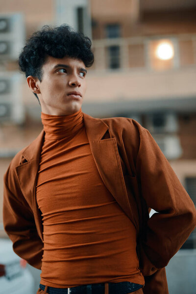 A stylish young man stands confidently outdoors, showcasing a modern orange outfit against a blurred urban backdrop
