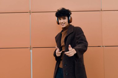 Kulaklıklı, turuncu bir duvarın önünde kameraya baş parmak işareti yapan şık bir genç adamın moda fotoğrafı..