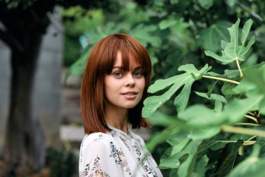 Canlı kızıl saçlı kadın yemyeşil bahçede poz veriyor. Bitkiler ve çalılarla birlikte.