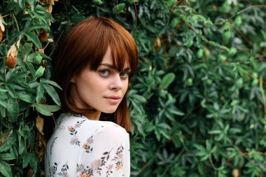 Canlı kızıl saçlı kadın yemyeşil bitki duvarının önünde duruyor.