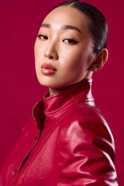 Kırmızı deri ceketli Asyalı esmer kadın elleri kalçasında pembe arka plana bakıyor. moda konsepti.