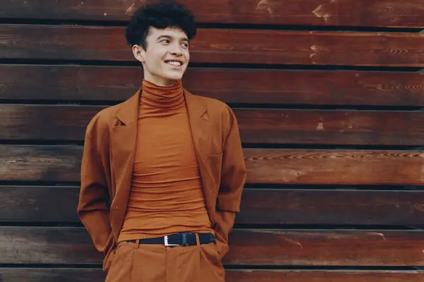 stock image A cheerful young man dressed in a brown turtleneck and jacket, standing relaxed against a rustic wooden wall, showcasing a contemporary fashion style