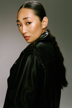 Asyalı kadın, siyah deri ceketli, gri arka planda poz veriyor. Asyalı genç bir kadının şık ve şık portresi..