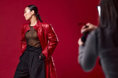 Kırmızı deri ceketli ve siyah pantolonlu Asyalı bir kadın kırmızı arka planda fotoğraf çekiyor. Moda tutkunları şık bir görünüş yakalıyor..