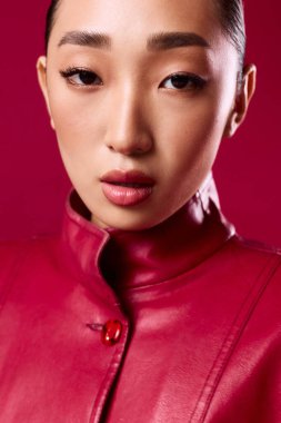 Kırmızı deri ceketli Asyalı esmer, kırmızı arka plandaki kameraya bakıyor. Şık görünümlü güzel bir kadın..