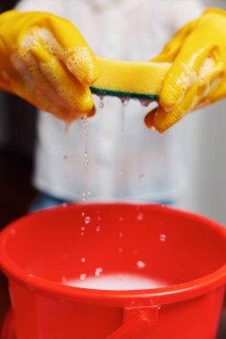 Sarı lastik eldiven takan bir kadın kırmızı kovanın üzerinde sünger tutuyor. Suyla dolu. Üstüne de başka bir sünger takılmış. bir temizlik konsepti resmi.