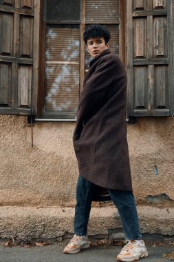 Kahverengi ceketli ve mavi kotlu şık bir adam. Binanın önünde duruyor. Elleri kalçasında, spor ayakkabıları giyiyor..