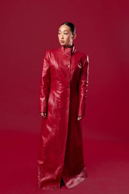 Kırmızı deri ceketli Asyalı esmer, elleri kalçasında ve başı yana eğilmiş kırmızı arka planda duruyor..