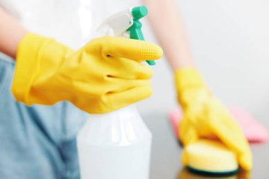 Sarı lastik eldivenli bir kadın elinde bir şişe temizlik solüsyonu ve masada bir sünger tutuyor..