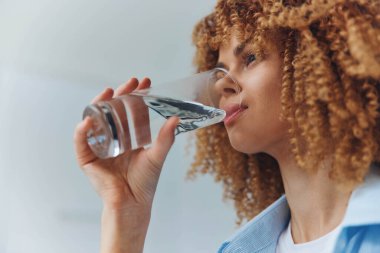 Sıcak bir odada kitap okurken cam şişeden su içen kıvırcık saçlı bir kadın.