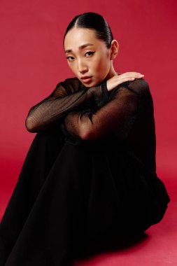 Siyah bluzlu ve pantolonlu Asyalı kadın elleri dizlerinin üstünde, kırmızı arka planda oturuyor. moda konsepti.