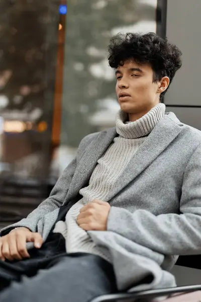 stock image Young man in grey sweater and black pants sitting on bench in front of building, looking away from camera. fashion concept.