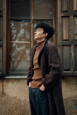 Kahverengi ceketli bir adamın, elleri ceplerinde pencerenin önünde dikilip yan tarafa bakarken moda fotoğrafı..