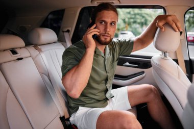 Düşünceli bir yüz ifadesi olan, telefonla konuşan genç bir adamın yer aldığı şık bir arabanın içi modern ulaşım ve iletişimi yakalıyor.