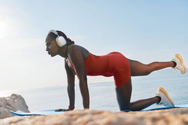 Kırmızı spor kıyafetli odaklanmış bir kadın okyanusun kenarındaki bir minderde yoga yapıyor ve sakin günbatımının tadını çıkarıyor.