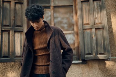 Kahverengi ceketli, şık bir genç adam eski bir binanın önünde poz veriyor. Penceresi kepenkli ve duvarları kahverengi..