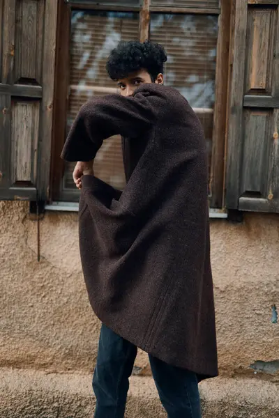 stock image Fashion man in a brown coat standing in front of a closed window with his hands on his hips and head tilted to the side.