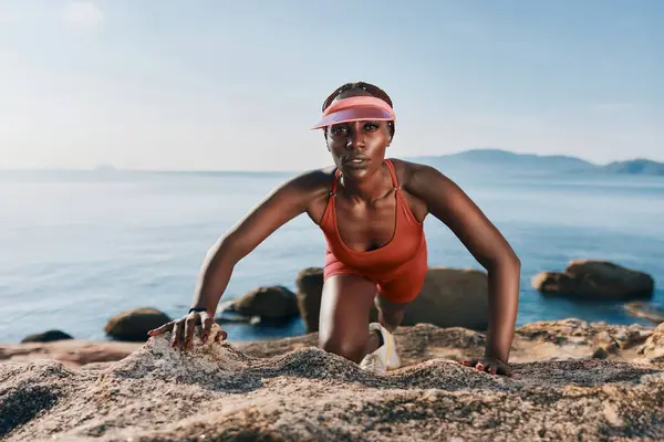 stock image A fit woman in a vibrant red outfit engaged in a climbing exercise on sandy rocks, against a serene ocean backdrop