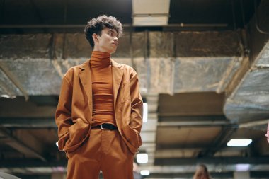 Havalı turuncu takım elbise ve balıkçı yaka giyen modaya uygun bir erkek model metalik detaylarla modern, endüstriyel bir yerde poz veriyor.