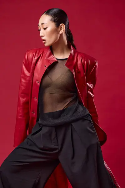 stock image Asian fashion model in red leather jacket and black pants posing with hands on hips and eyes closed on red background. trendy and stylish look.