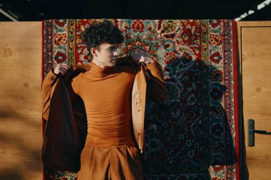 Turuncu balıkçı yaka ve pantolon giyen şık bir genç adam ahşap bir arka plana karşı renkli bir halının önünde güvenle duruyor.