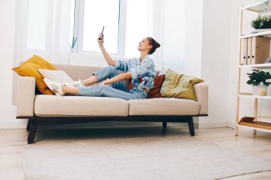 Mutlu kadın cep telefonu tutuyor ve rahat oturma odasında kanepede dinlenirken gülümsüyor.