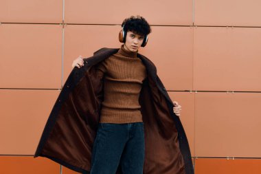 Kulaklıklı, kahverengi ceketli, yüzünde ve kalçasında bir gülümsemeyle turuncu bir duvarın önünde duran şık bir genç..