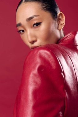 Kırmızı arka planda fotoğraf için poz veren kırmızı deri ceketli Asyalı esmer kadın..