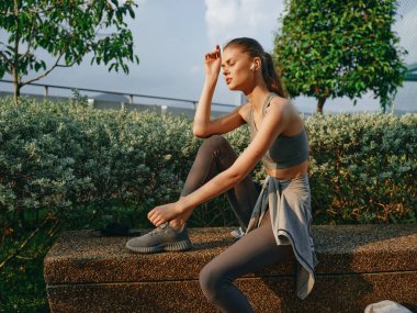 Formda bir kadın taş bir çıkıntıda oturur, sakin doğal bir zemine karşı spor kıyafetleriyle bacağını esnerdi.