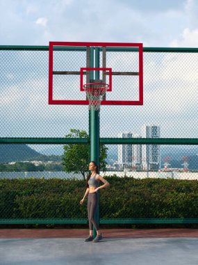 Spor kıyafetli genç bir kadın açık bir sahada basketbol potası ile poz veriyor, arkasında da bir şehir manzarası var.