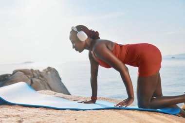 Formda bir kadın, kayalık bir plajda mavi bir hasırla yoga yapar, kulaklık takar, arka planda dingin deniz vardır. Parlak ve sakin bir atmosfer.