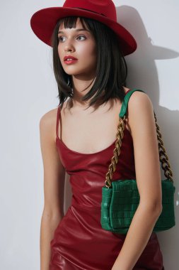 Şık kırmızı şapkalı ve elbiseli bir kadın, elinde yeşil bir çanta ve kırmızı bir çanta olan beyaz bir duvara karşı duruyor, cesur bir moda ve canlı bir kontrast hissi uyandırıyor.