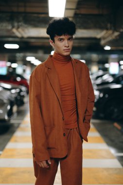 Kahverengi takım elbise ve balıkçı yaka giyen şık genç bir adam park etmiş arabalarla dolu bir otoparkta güven içinde poz veriyor.