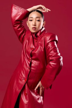 Kırmızı deri ceketli Asyalı esmer, elleri kalçasında kırmızı arka planda poz veriyor..