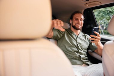 Neşeli genç bir adam arabanın arka koltuğunda oturuyor. Akıllı telefonunu video görüşmesi için kullanıyor. Etrafı bej renkli deri koltuklar ve yeşil yapraklarla çevrili.