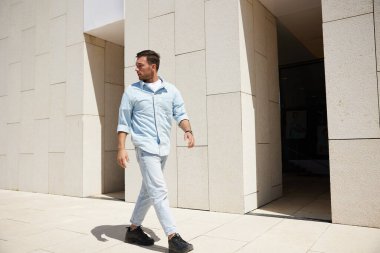 Kendine güveni tam genç bir adam modern bir binanın önünden geçiyor ve yan tarafa bakıyor. Görüntü güven ve kentsel yaşam tarzını aktarıyor..