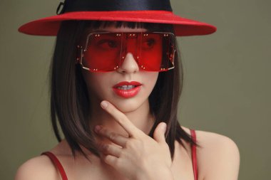 Kırmızı şapkalı ve güneş gözlüklü kadın, elleri çenesinde ve parmakları dudaklarında poz veriyor, şık ve düşünceli bir ifade sergiliyor, moda veya düşünsel temalı görseller için mükemmel.