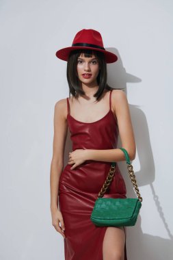 Kırmızı elbiseli, kırmızı şapkalı, yeşil çantalı, kırmızı çantalı, beyaz çantalı hayat dolu kırmızı elbiseli ve uyumlu şapkalı şık bir kadın temiz beyaz arka plana karşı yeşil çanta ve kırmızı çanta taşıyor.