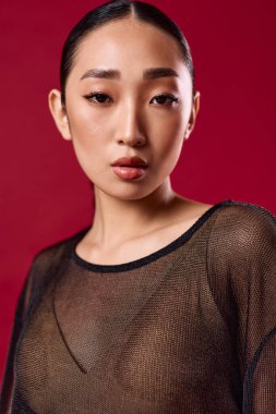 Asyalı bir manken kırmızı arka planda poz veriyor. Elleri kalçasında, gözleri yana bakıyor..