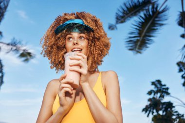 Neşeli genç bir kadın açık mavi bir gökyüzünün altında gülümseyen meyve kokteyli tutuyor. Güneşlik takıyor ve etrafı tropikal palmiye ağaçlarıyla çevrili.