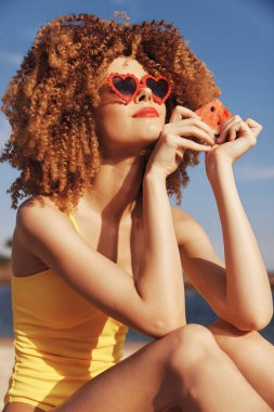 Sarı mayo giymiş şık bir kadın bir dilim karpuz ile poz veriyor.