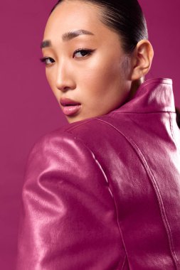 Pembe deri ceketli Asyalı esmer, mor arka planda poz veriyor, at kuyruklu saçlı, gözleri kapalı..