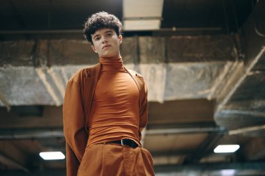 Tarz sahibi bir erkek model turuncu bir balıkçı yaka giyer ve uyumlu pantolonlar giyer. Endüstriyel arka planda modern moda sergilerler.